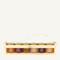6 jars of honey, 50 g each: blossom honey and five flavoured honeys in a wooden gift crate with a rope handle, on a warm light background.