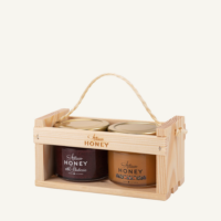 Two 100 g jars of blossom honey and honey with blueberries in a wooden gift crate with a rope handle, on a warm light background.
