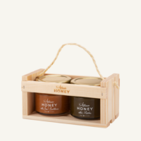 Two 100 g jars of honey with sea buckthorn and honey with matcha in a wooden gift crate with a rope handle, on a warm light background.