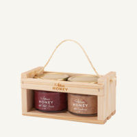 Two 100 g jars of honey with cranberries and honey with wild berries in a wooden gift crate with a rope handle, on a warm light background.