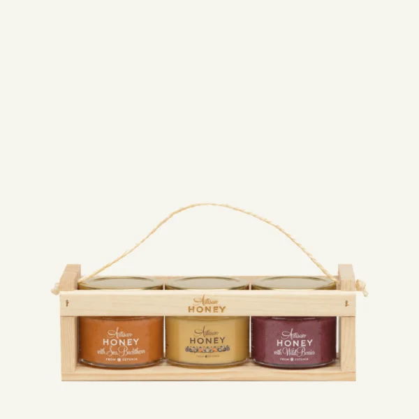 Three 100 g honey jars in a wooden crate: sea buckthorn, blossom honey and wild berries, on a warm light background.